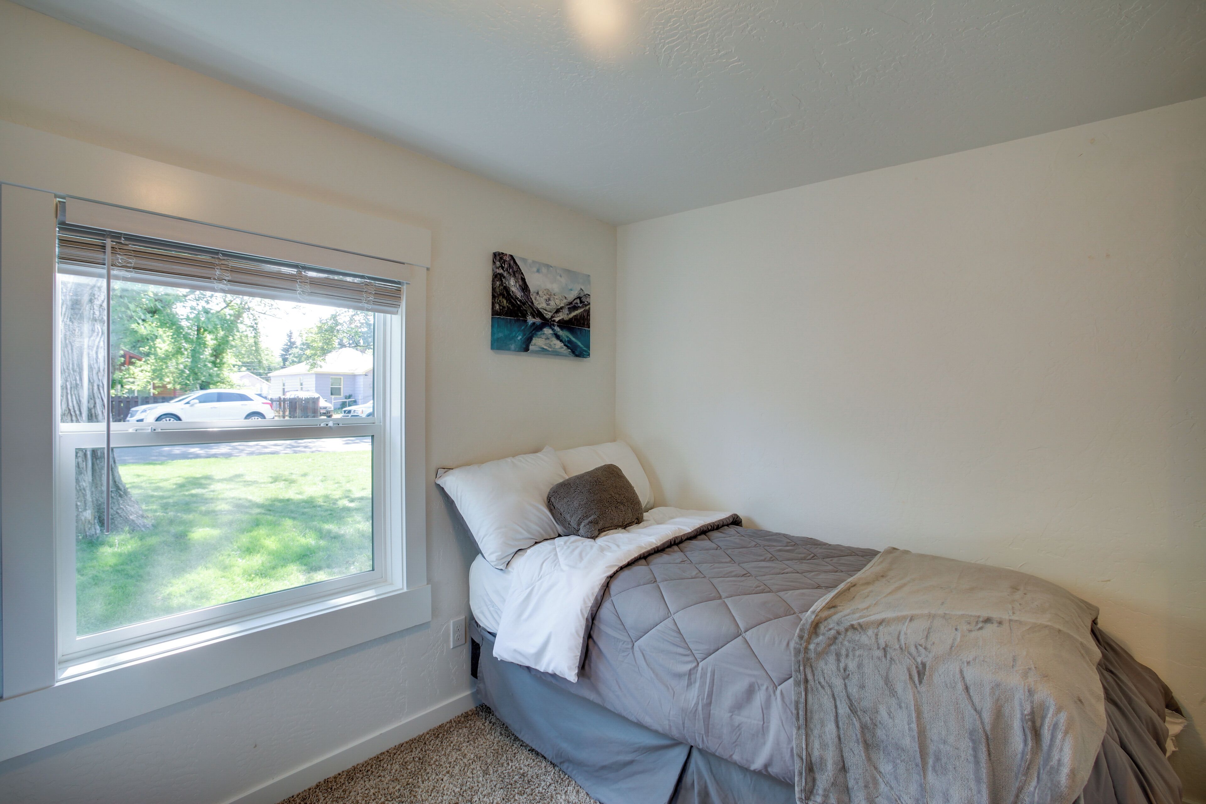A bedroom with a bed, a window with blinds, a painting on the wall, and a view of a parking lot and trees outside.