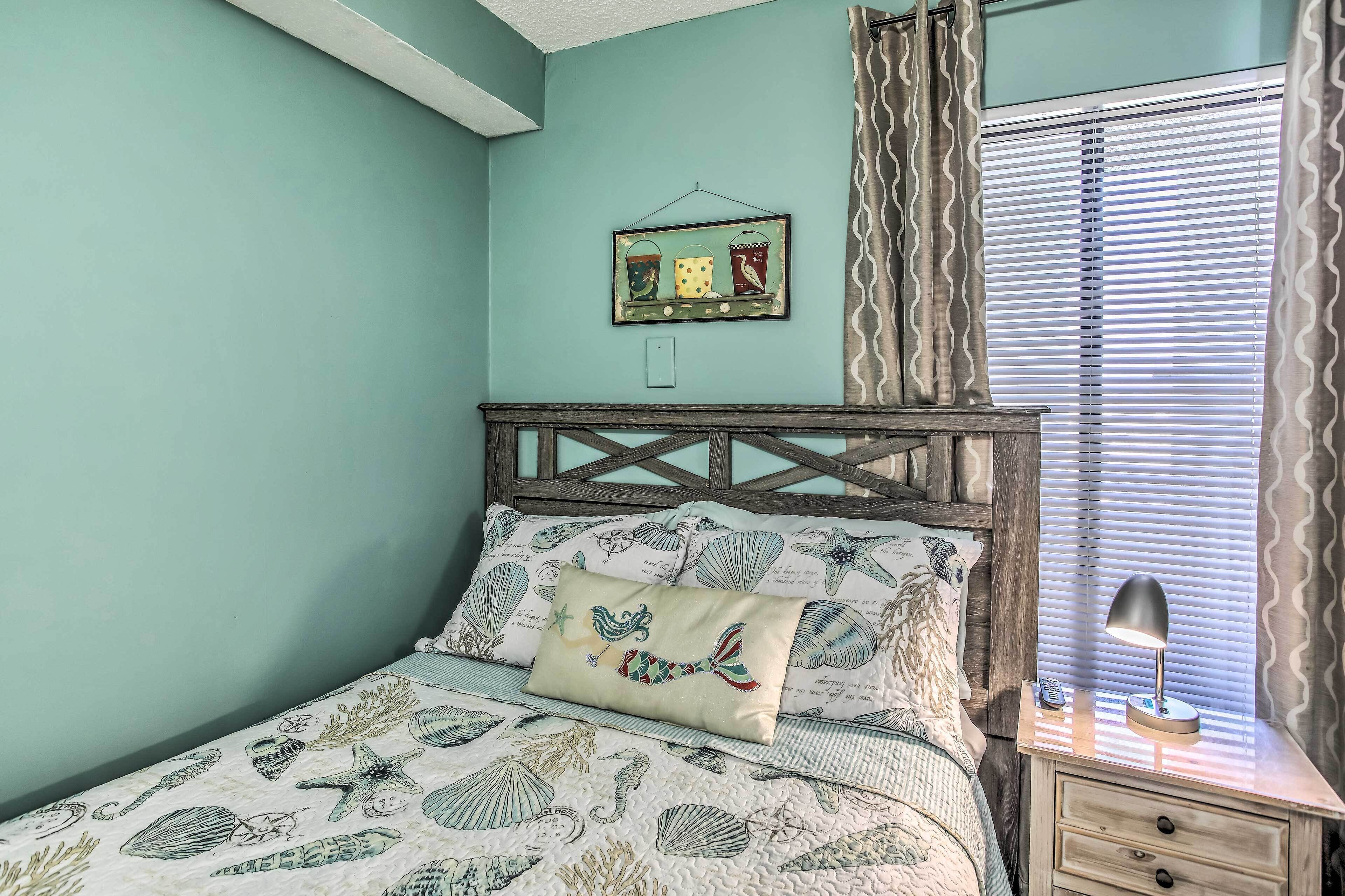 A bedroom with a bed featuring a sea-themed bedspread, a wooden headboard, a bedside table with a lamp, and a window with blinds.