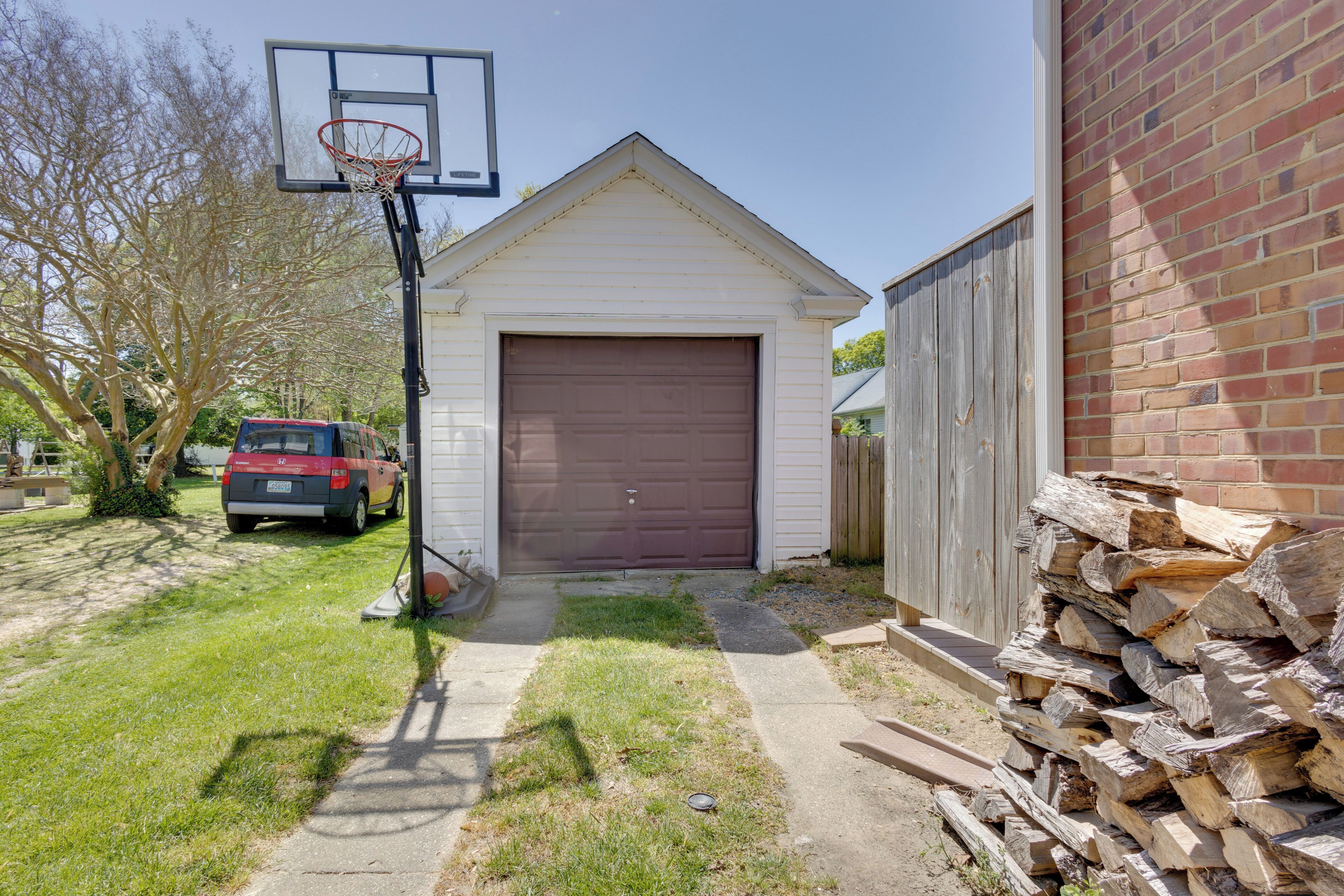 A garage with a basketball hoop, a red vehicle parked outside, and a stack of firewood.