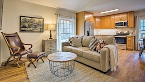 Amplia sala de estar con sofá, sillón y mesa de centro. Se ve una cocina con armarios de madera y electrodomésticos al fondo.