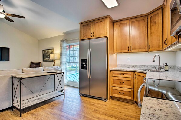 A modern kitchen with wooden cabinets, a stainless steel refrigerator, a granite countertop, and a window with blinds. - Walk to Main Street Eats: Remodeled Saluda Apt (Saluda)