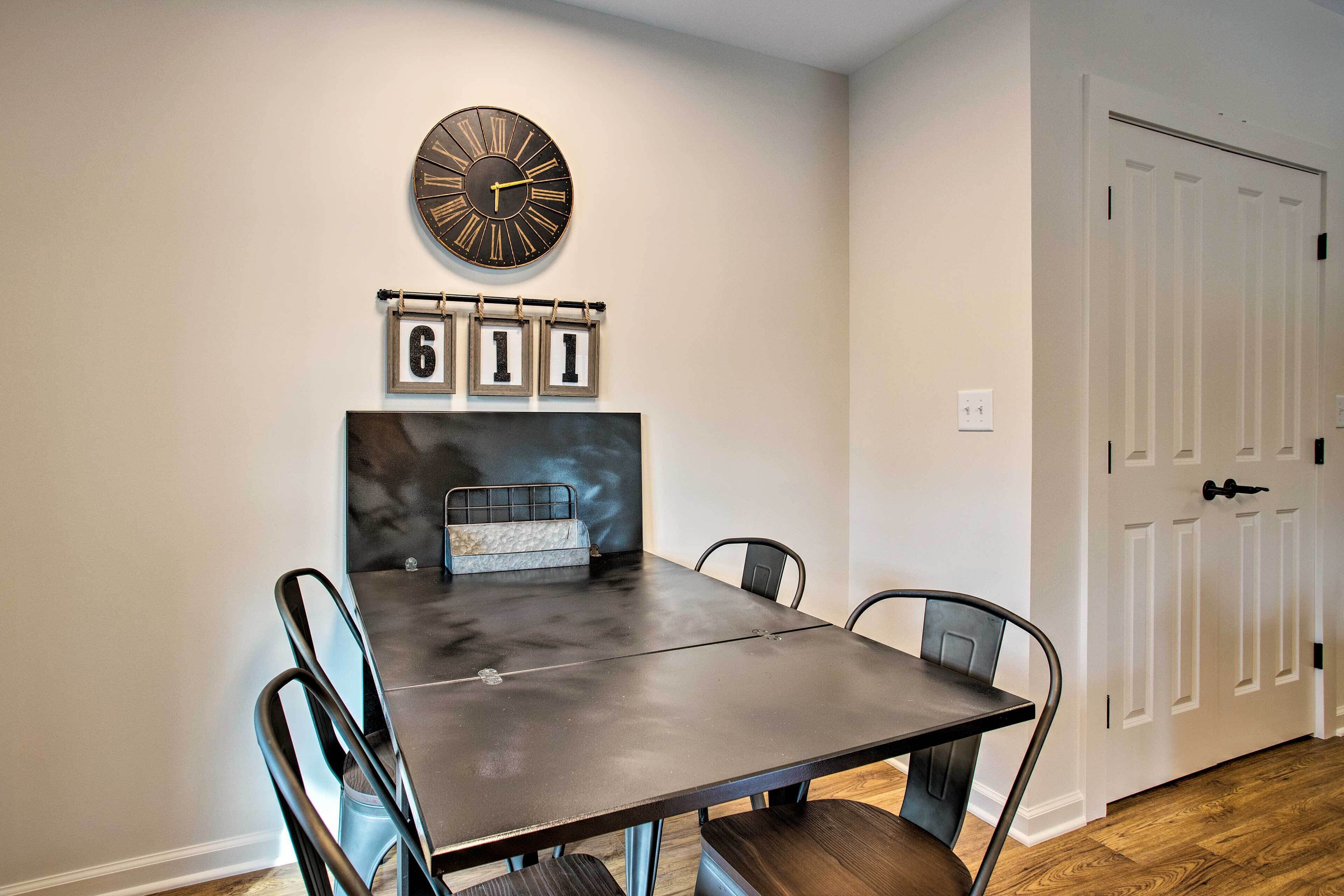 Un comedor con una mesa de madera oscura, cuatro sillas, un reloj y un panel con números enmarcados en la pared.