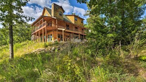 A large wooden cabin with multiple decks and a prominent chimney, surrounded by dense forest. - Spacious Log Cabin w/ Fire Pit & Game Room! (Eureka Springs)
