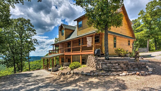 A large log cabin with a porch, a stone staircase, and a view of the mountains. - Spacious Log Cabin w/ Fire Pit & Game Room! (Eureka Springs)