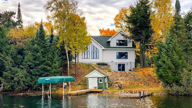 A two-story house with a dock extending into a lake, surrounded by autumn-colored trees. - Fishing Haven: Family Home on Indian Lake (Rhinelander)
