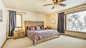 A bedroom with a large bed, a ceiling fan, a nightstand, a window with curtains, and a view of trees outside. - Fishing Haven: Family Home on Indian Lake (Rhinelander)