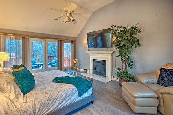 A bedroom with a large bed, a fireplace, a TV mounted above it, and a balcony with outdoor furniture.