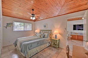 A bedroom with a wooden ceiling, a bed with a headboard, two bedside lamps, a TV, and a small dining area with a table and chairs.