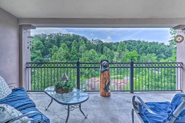 A balcony with a view of a forest, a wooden sculpture, and a glass table with a plant.