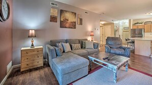 A living room with a sofa, ottoman, coffee table, and a side table with lamps. There is a kitchen area visible in the background. - Hunter Mountain Ski Retreat - Steps to Lift! (Hunter)