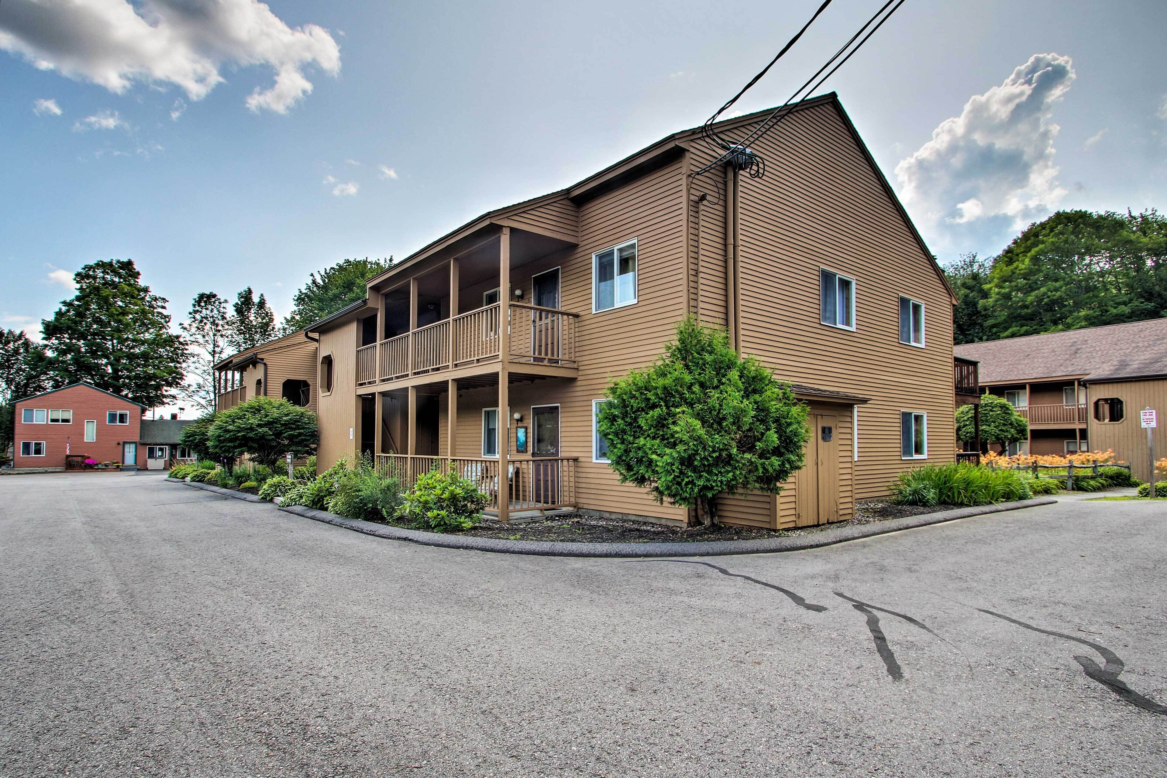 A two-story building with a balcony on the second floor, surrounded by trees and a gravel driveway.