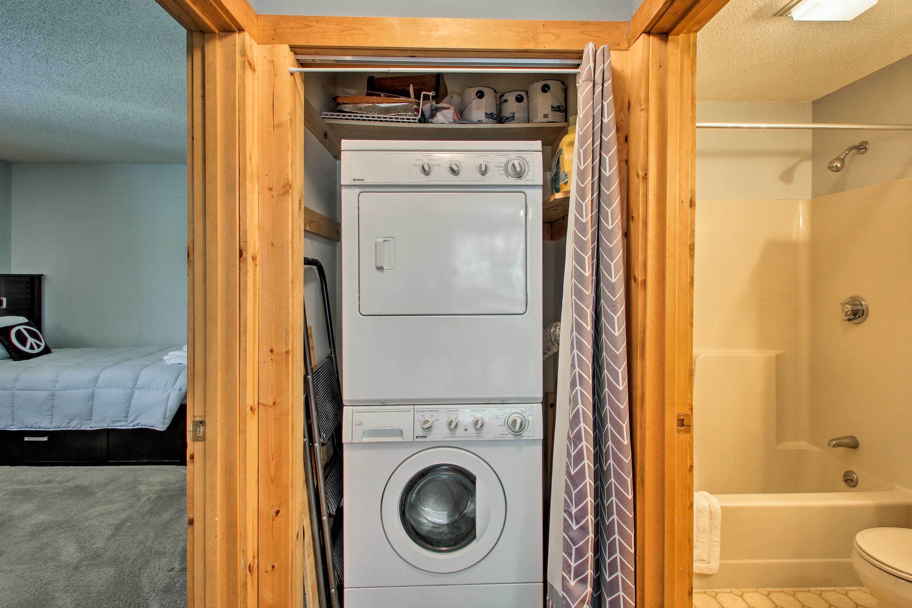 A compact laundry room with a stacked washer and dryer, a shower, and a bed with a blue blanket in the adjacent room.