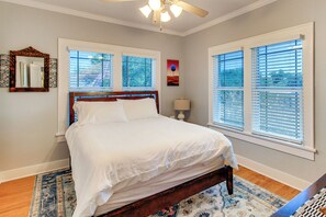 Quarto com cama, duas janelas com persianas, ventilador de teto, mesa de cabeceira e tapete.