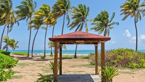 Ein Strandpavillon mit Holzdach und offenen Seiten, umgeben von Palmen und freiem Blick auf das Meer.