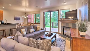 Ein modernes Wohnzimmer mit einer Couch, einem Couchtisch und einem Fernseher. Ein Kamin und ein großes Fenster mit Blick auf Grünflächen sind ebenfalls vorhanden.