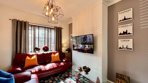 A living room with a red leather sofa, a colorful patterned rug, a glass coffee table, and a wall-mounted flat-screen TV displaying a travel channel.