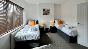 A hotel room with two beds, a radiator, a window with blinds, a wall-mounted light fixture, and a door.