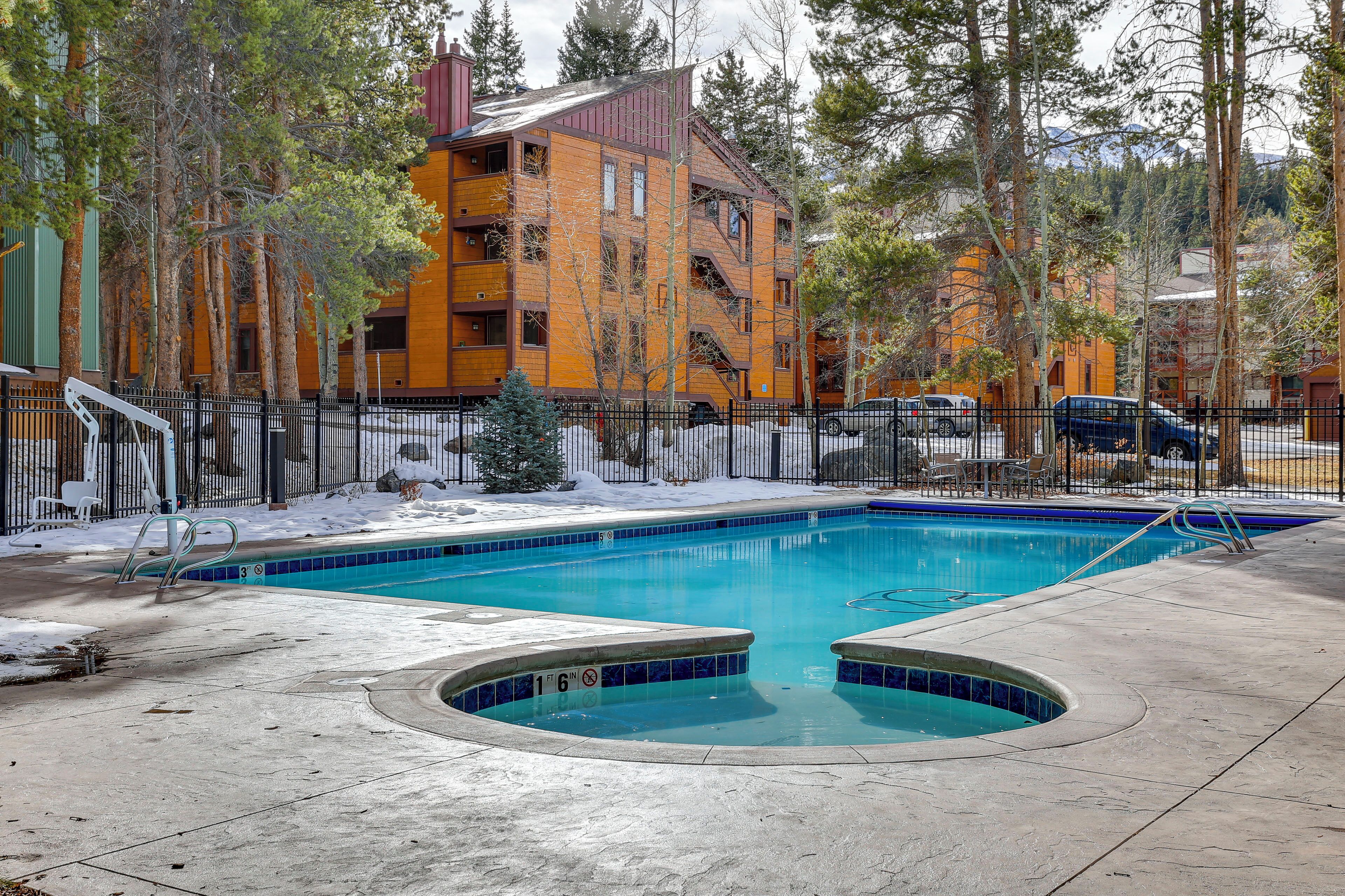 Una piscina con un deck perimetral, un pequeño árbol de Navidad y un edificio de varios pisos con balcones al fondo.