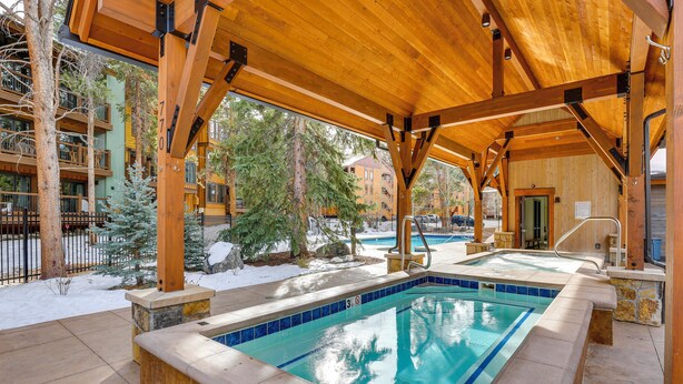 En utomhuspool med tak, omgiven av träd och en byggnad med balkonger, och med ett trädäck runt poolen.
