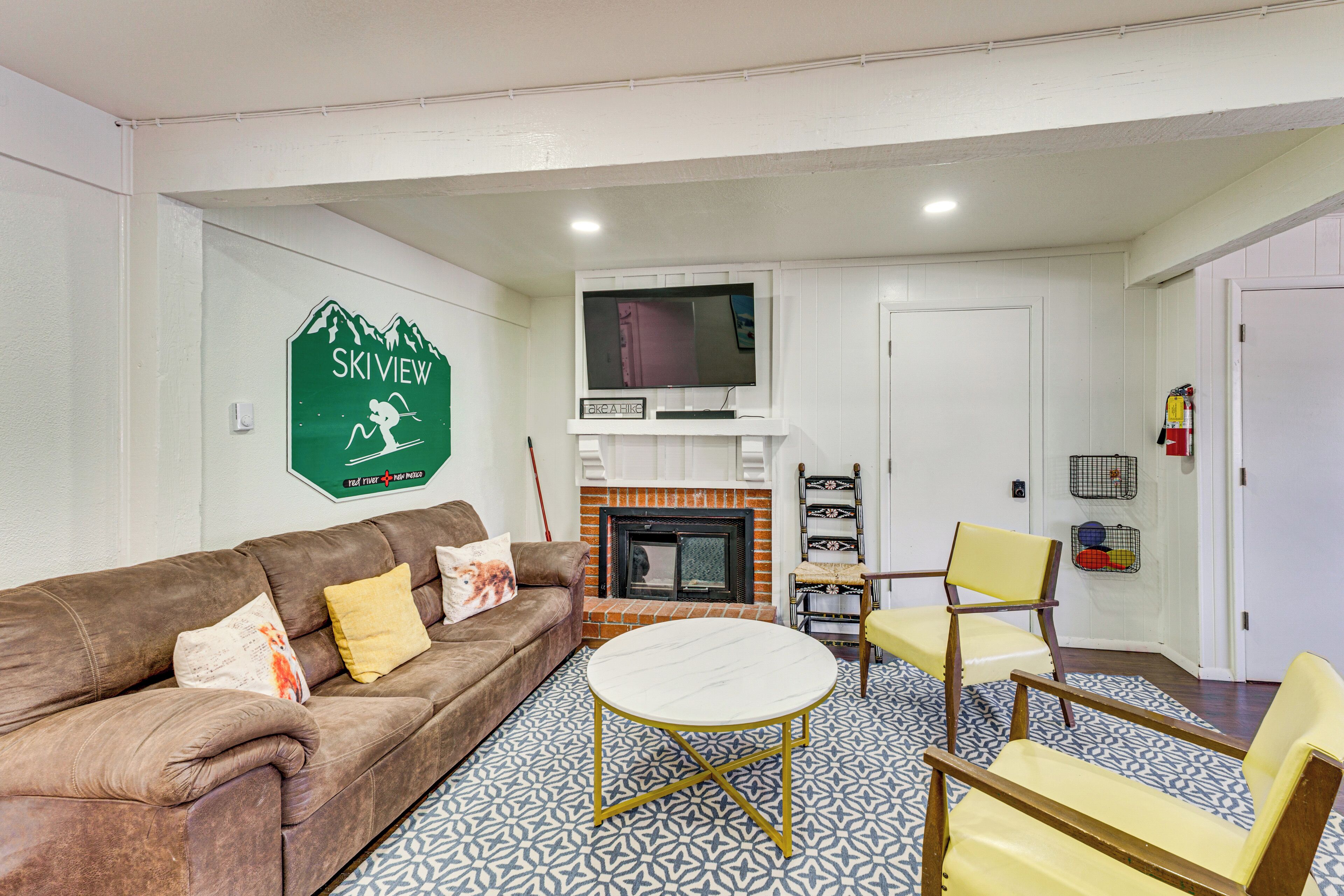 A living room with a brown leather sofa, a round white table, and two yellow chairs. A television is mounted above a fireplace with a red brick surround.