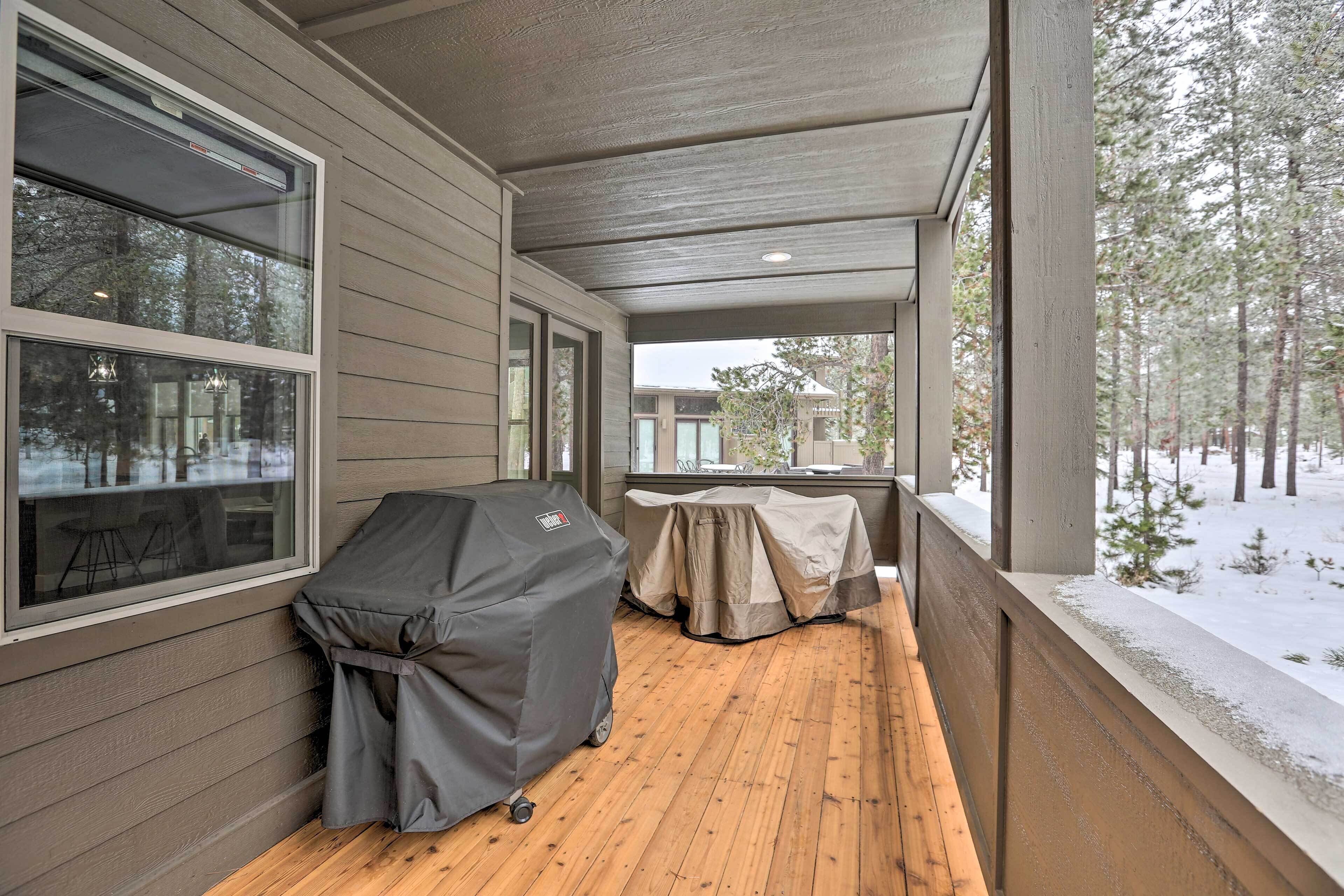 A covered porch with a grill and table, wooden flooring, and a view of a snowy landscape.