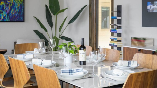Une table à manger est dressée avec de l’assiette blanche, des serviettes et des verres à vin, et entourée de chaises en bois. Une bouteille de vin est posée sur la table.