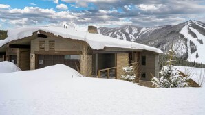 Une cabane en bois avec un garage, entourée de montagnes enneigées et sous un ciel nuageux.
