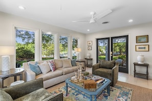 A spacious living room with a beige sofa, a blue coffee table, and a view of a garden through large windows. - Live Oaks & Marina Views! Ocean Springs Home (Ocean Springs)