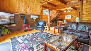 L’intérieur chaleureux d’un chalet en bois présente un plafond en bois, un canapé en cuir, une table basse, un téléviseur et un escalier menant à un étage supérieur.
