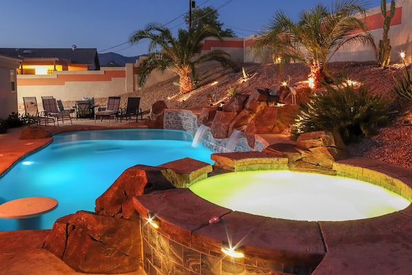 Relax by the serene pool surrounded by natural rock formations and palm trees, with lounge chairs and a table for a peaceful retreat.