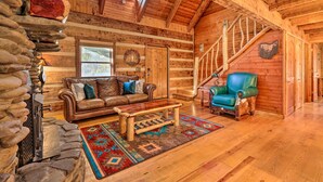 A log cabin interior with a stone fireplace, leather sofa, and a blue armchair. - Luxe Dog-friendly Cabin w/ Hot Tub & Game Room (Logan)