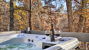 A hot tub with two glasses of red wine and a bottle, surrounded by trees with autumn foliage. - Luxe Dog-friendly Cabin w/ Hot Tub & Game Room (Logan)