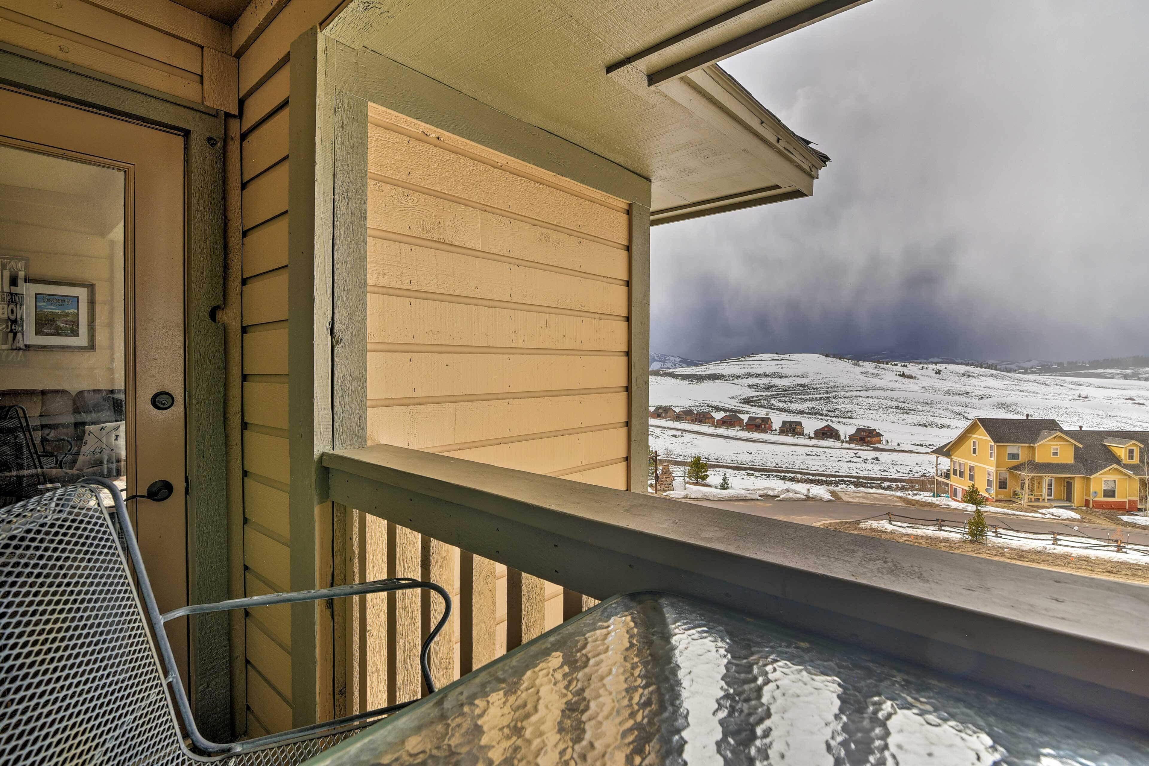 Un balcon avec une chaise en métal, une fenêtre donnant sur un paysage enneigé avec des maisons, et une porte ornée d’un cadre de photo.