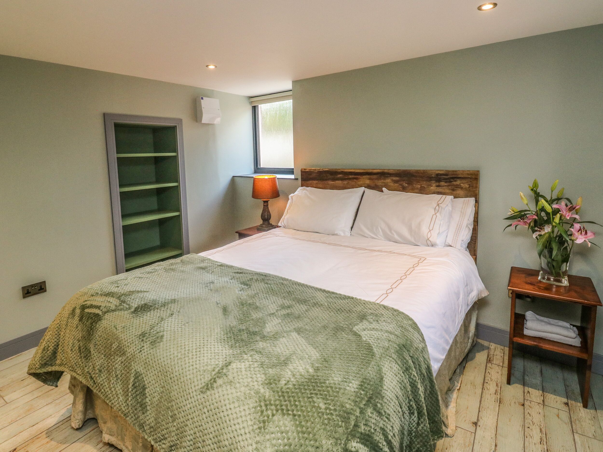 Un dormitorio con una cama grande, una mesita de noche con lámpara, un cabecero de madera, una manta verde de punto, suelo de madera, una ventana y un aire acondicionado de pared.