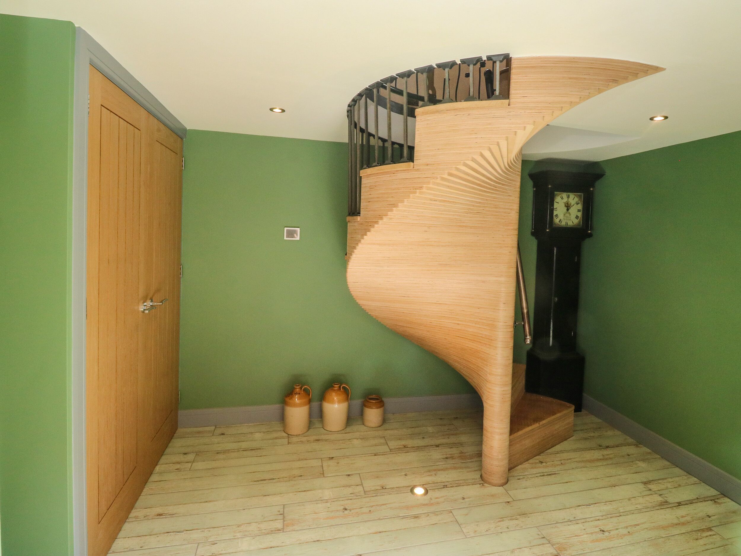 Una escalera de caracol con escalones de madera y barandilla negra, que lleva a una plataforma con un reloj y tres jarrones de cerámica en el suelo.