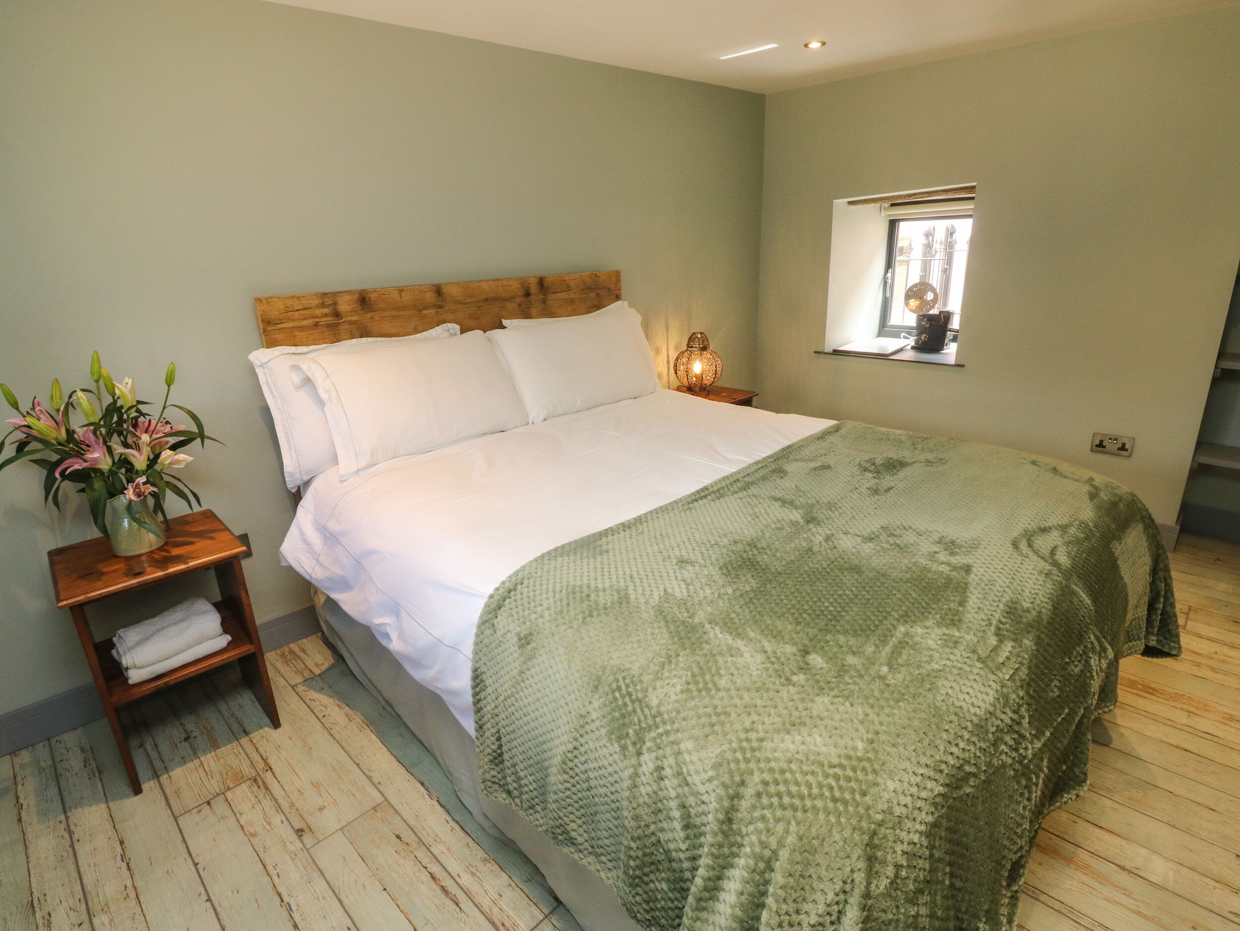 A bedroom with a wooden headboard, white bedding, a green blanket, a bedside table with a lamp, and a window with a view.