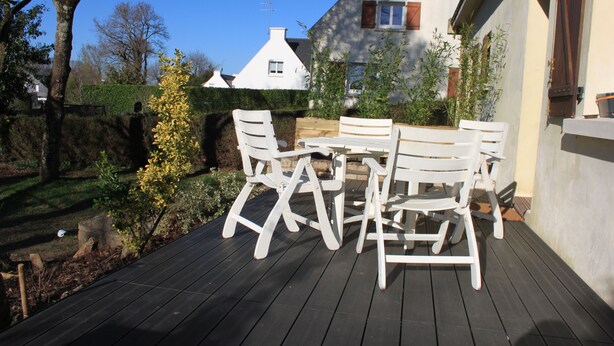 Une terrasse en bois, agrémentée de meubles d’extérieur blancs, entourée d’un jardin et d’une maison blanche au toit brun.