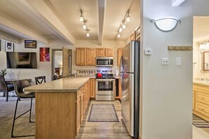 A modern kitchen with wooden cabinets, stainless steel appliances, a dining area with a table and chairs, and a flat-screen TV mounted on the wall. - Lakefront Condo w/ Mtn Views in Dillon! (Dillon)