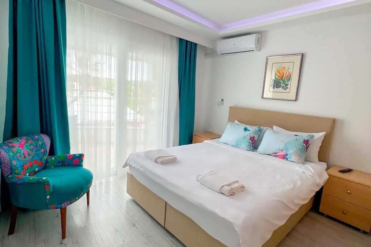 A hotel room with a bed, a chair, a bedside table, and an air conditioning unit.