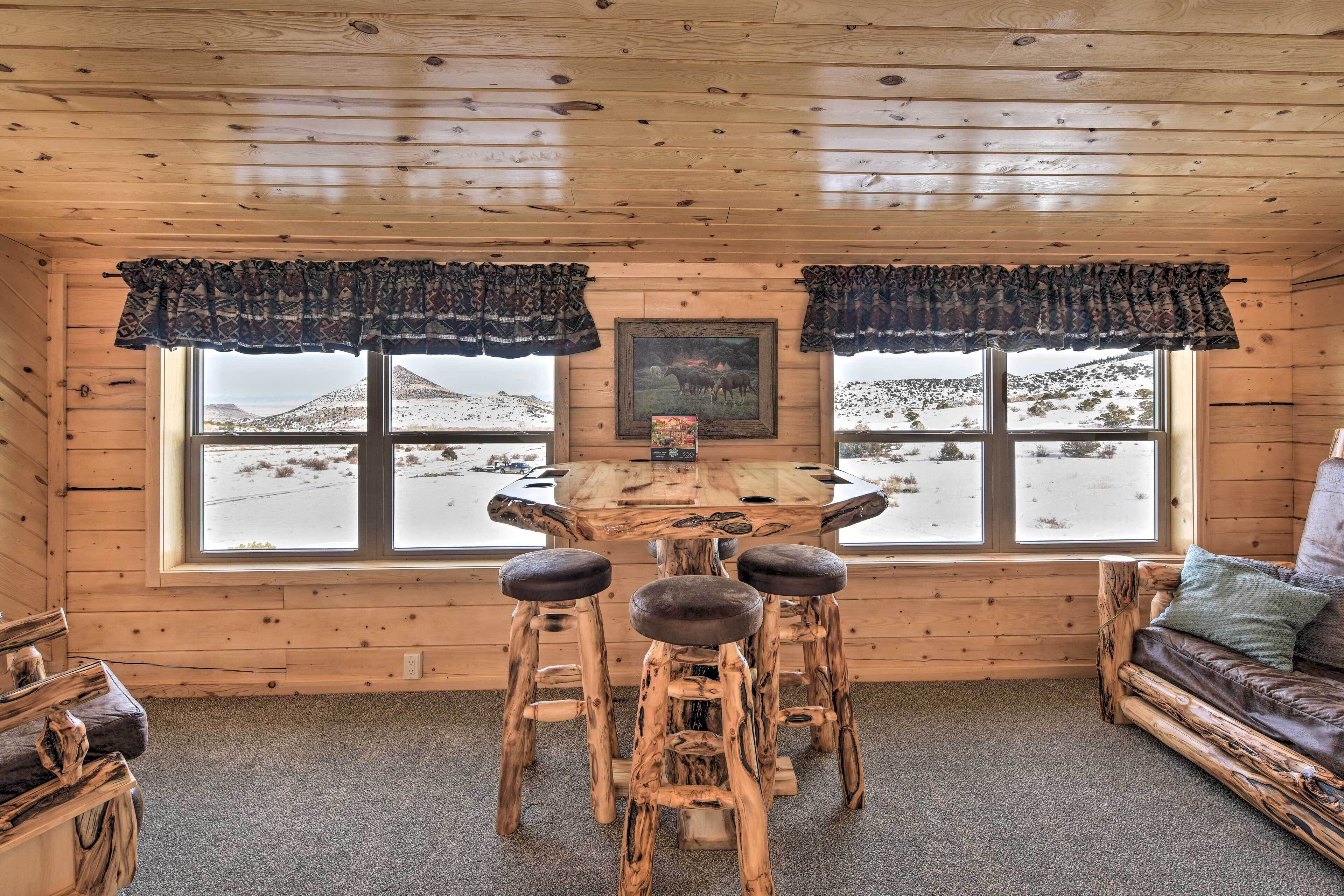 A rustic wooden interior with a round table and four stools, large windows with curtains, a framed picture on the wall, and a leather sofa.