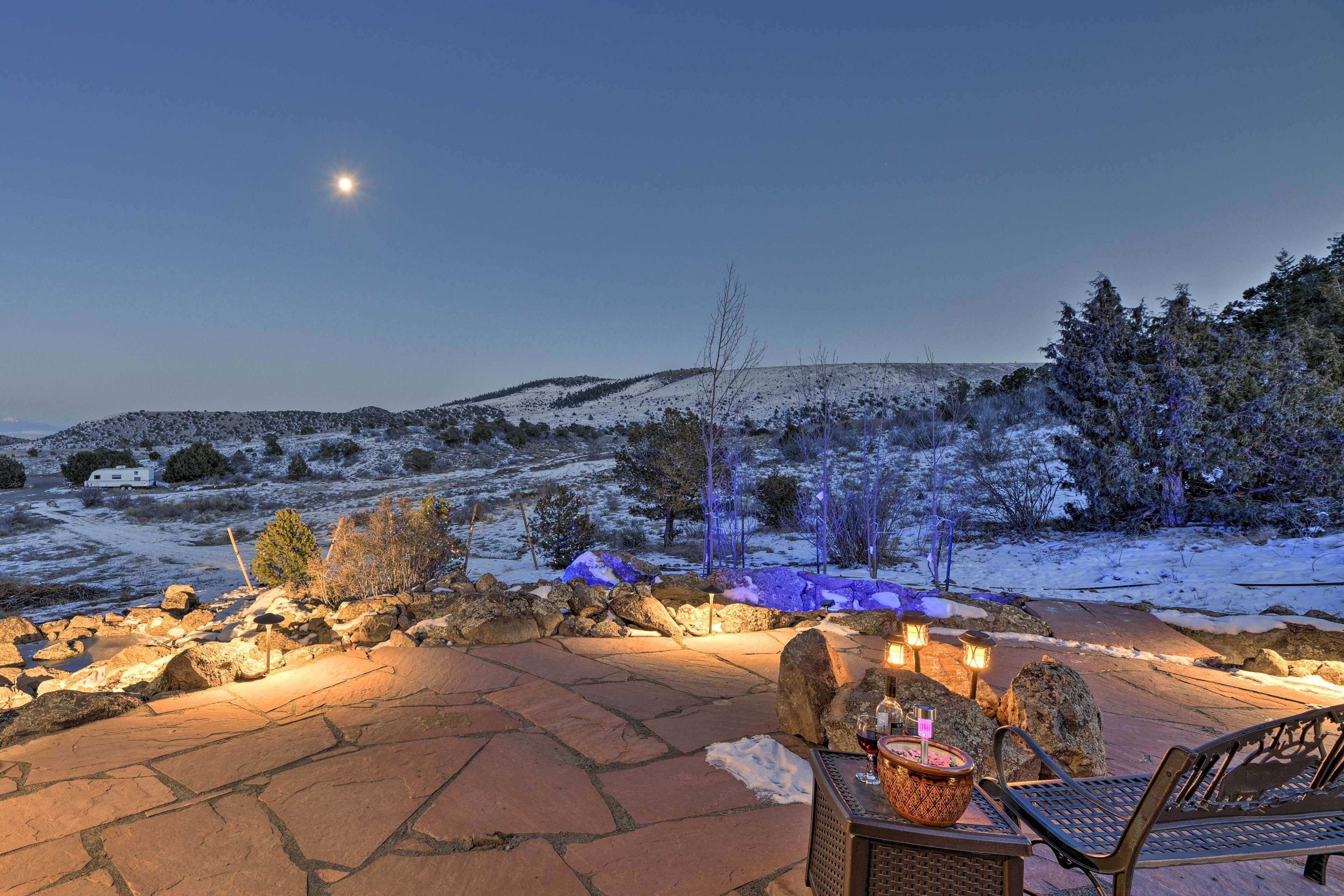 A patio with stone flooring, a metal bench, a table with a candle, and a view of snow-covered hills under a night sky.