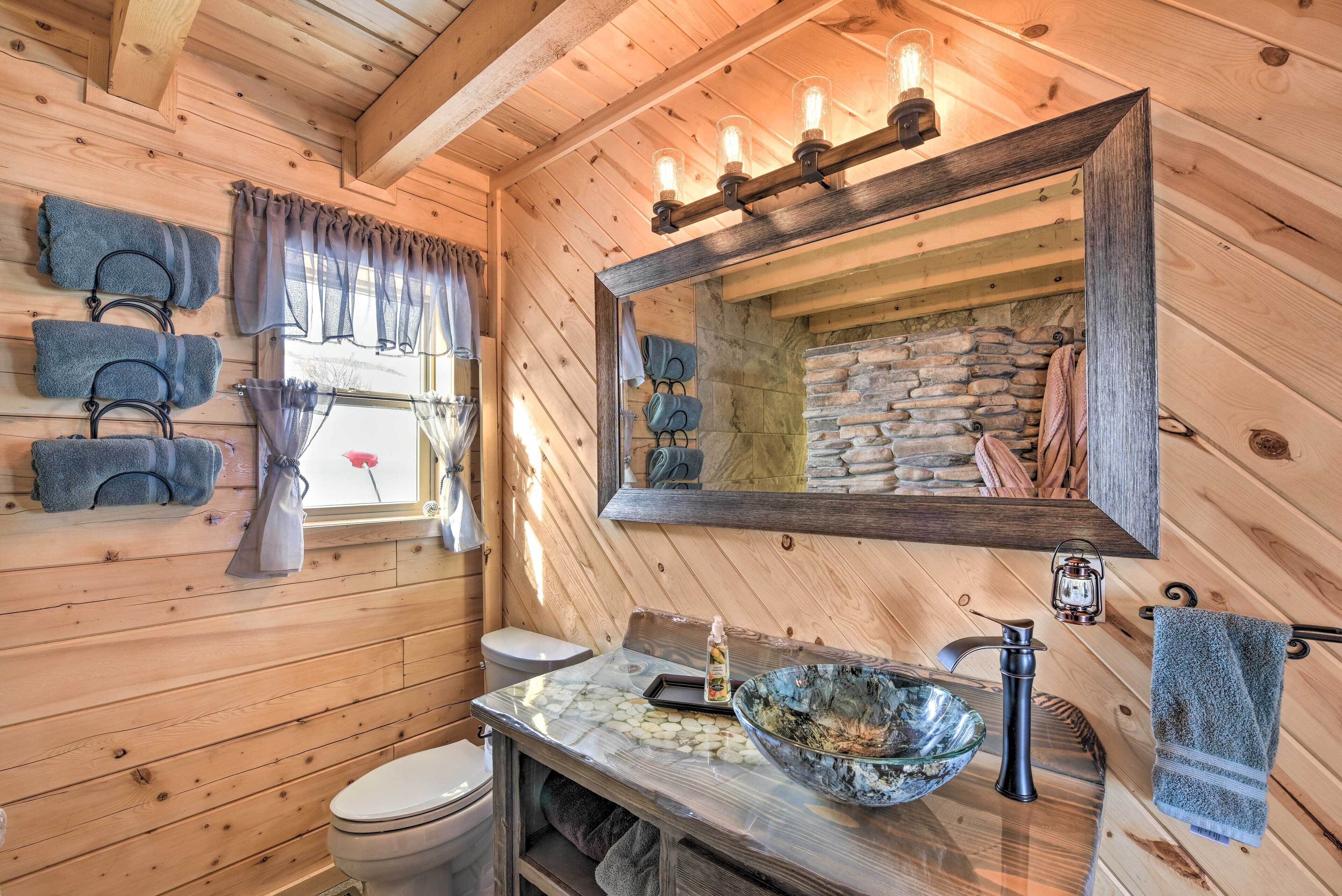 A wooden cabin bathroom with a stone wall, a wooden vanity, a large mirror, and a hanging towel rack.