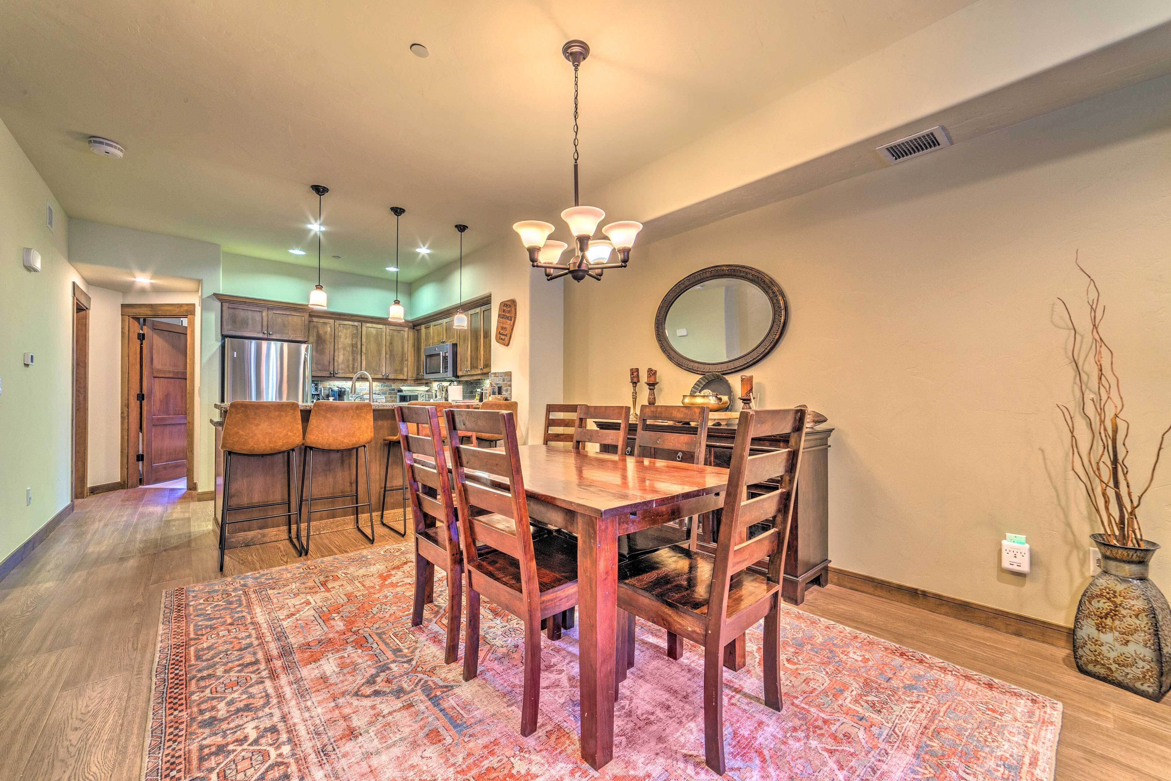 A dining area with a wooden table and chairs, a chandelier, and a kitchen in the background.