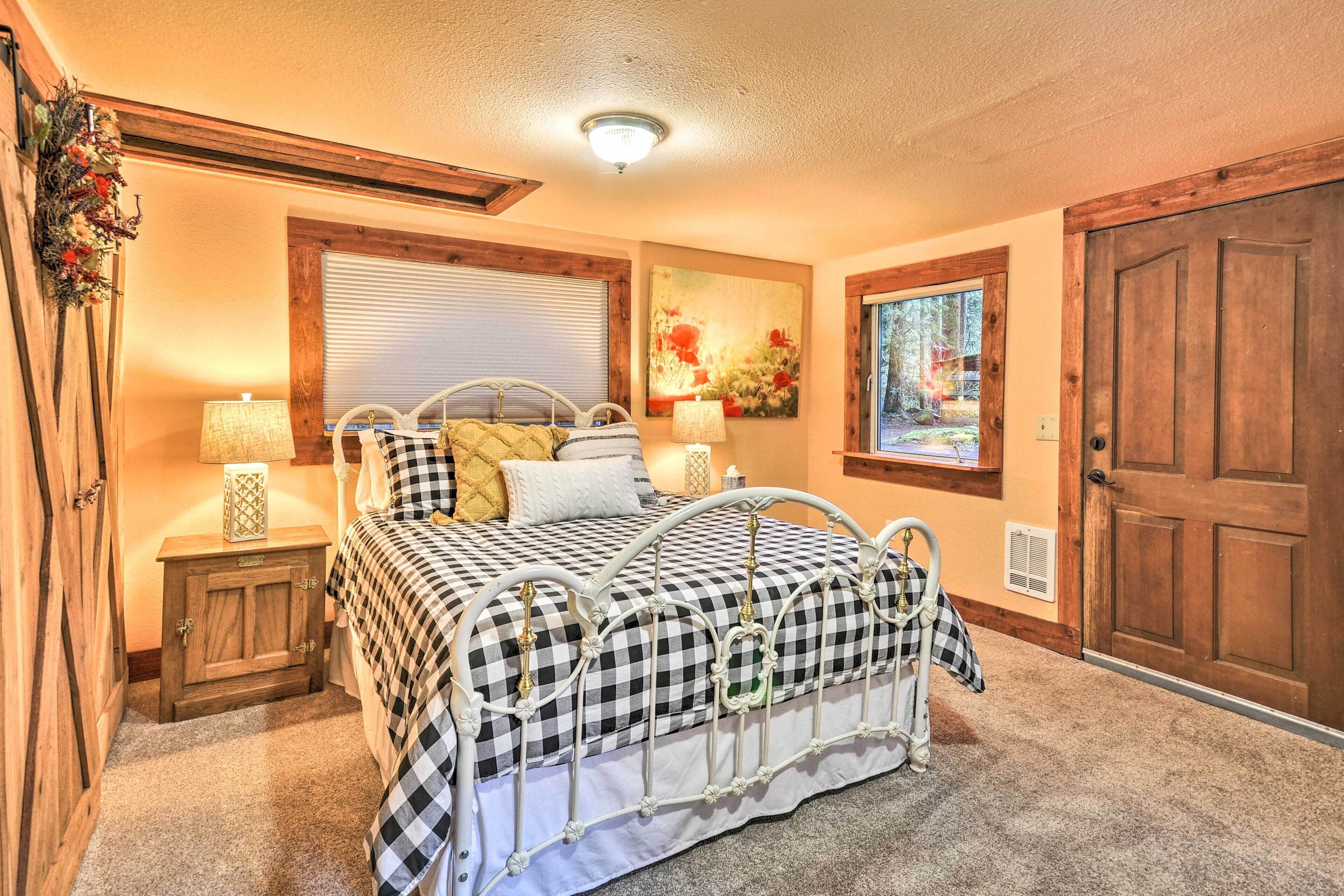A bedroom with a checkered bedspread, wooden headboard, two bedside tables with lamps, a window with blinds, a door, and a wall-mounted artwork.
