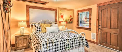 A bedroom with a checkered bedspread, wooden headboard, two bedside tables with lamps, a window with blinds, a door, and a wall-mounted artwork.