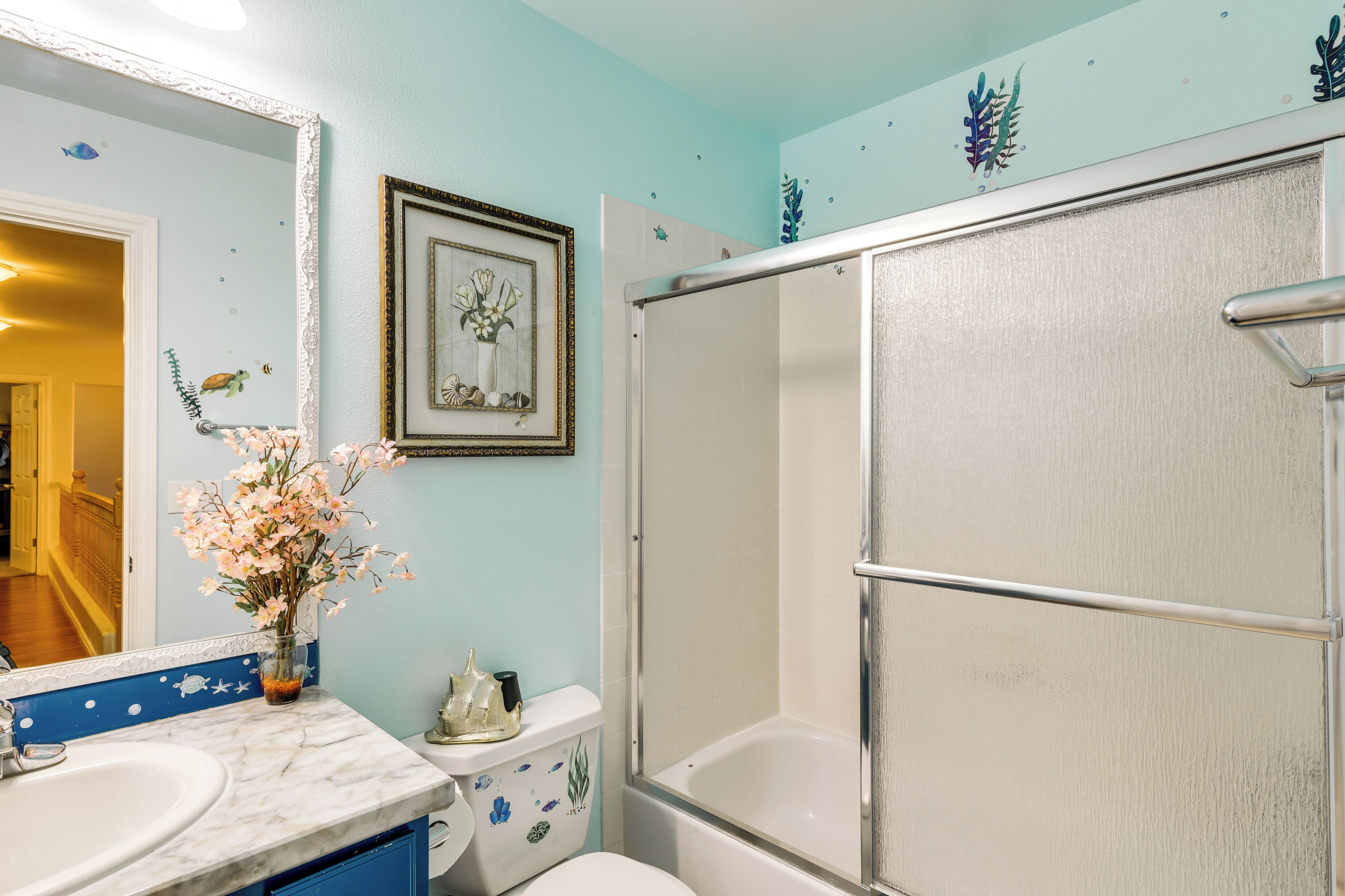 Une salle de bain avec un lavabo blanc, des toilettes et une douche. Un miroir est placé au-dessus du lavabo et un tableau encadré orne le mur.