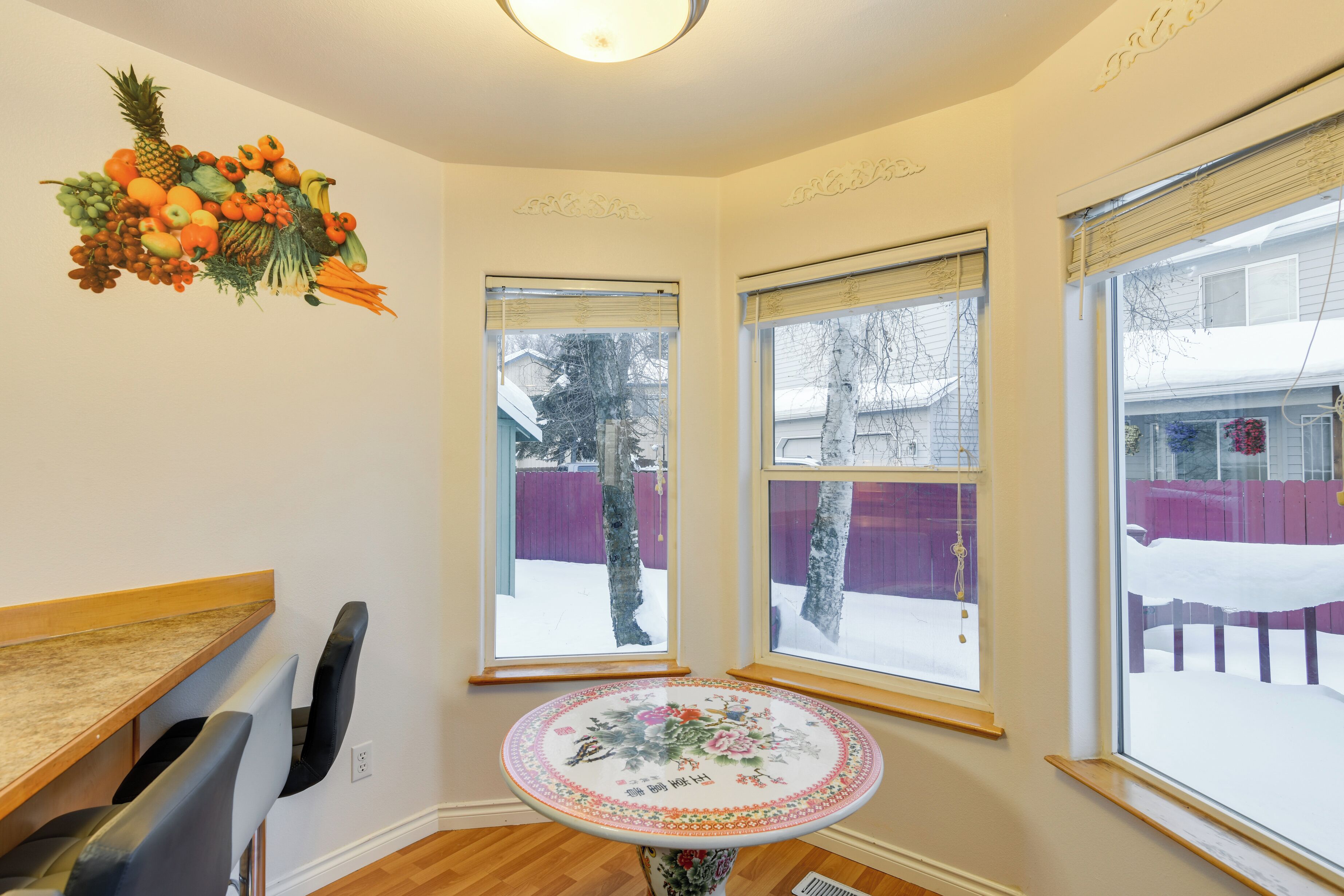A corner room with a dining table, chairs, a small decorative table, and three windows with a view of a snowy yard.