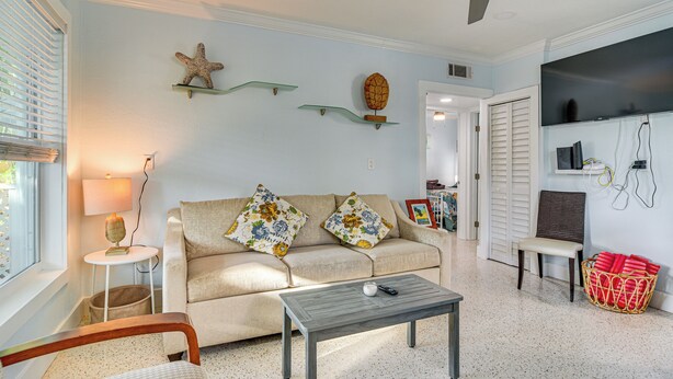 A living room with a sofa, a coffee table, a ceiling fan, a TV, and a wall-mounted shelf with decorative items. - Indian Rocks Beach Retreat, Walk to Gulf & Beaches (Indian Rocks Beach)