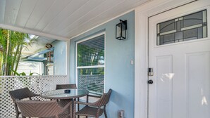 A covered patio with a round table and wicker chairs, a white door with a window, and a white fence. - Indian Rocks Beach Retreat, Walk to Gulf & Beaches (Indian Rocks Beach)
