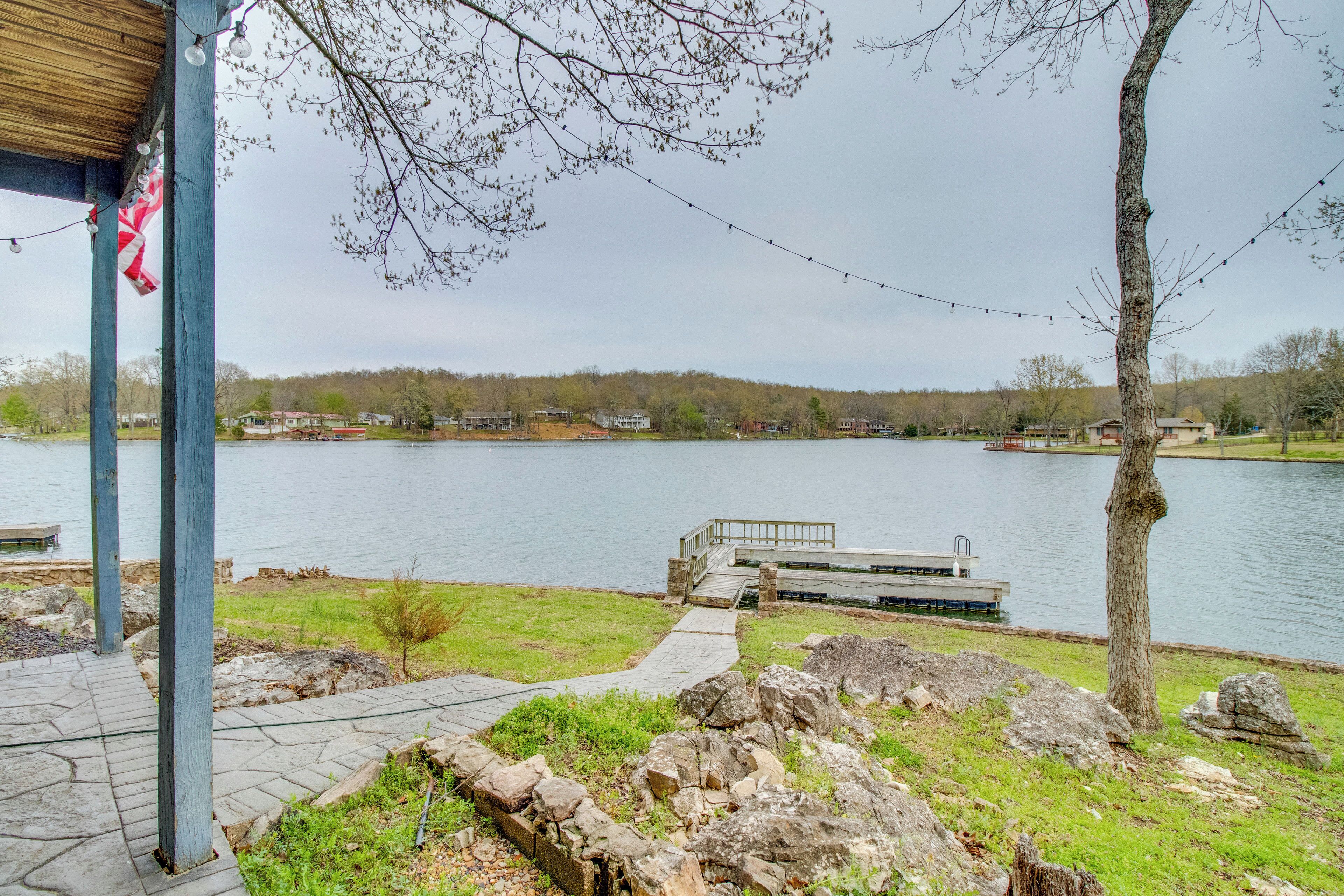 A lakeside walkway with a stone path, a wooden deck, and a view of the lake and distant houses.